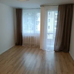 Jasne, puste pomieszczenie z panelami podłogowymi w odcieniach brązu, okno z białymi i brązowymi zasłonami oraz nowoczesną lampą sufitową z metalową konstrukcją.