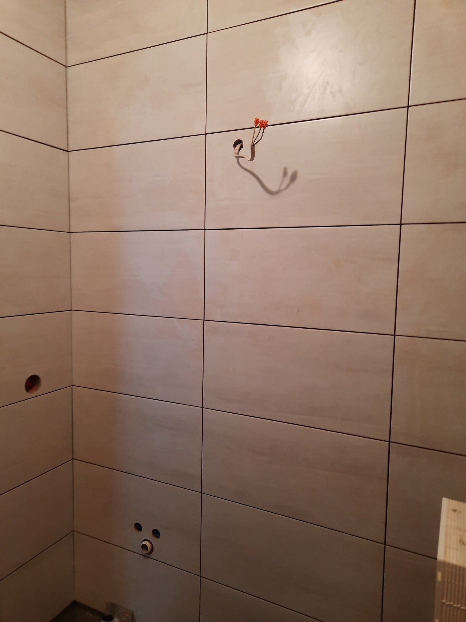 Fragment łazienki w trakcie prac: beżowe płytki ścienne, widoczne otwory na instalacje, wystające przewody elektryczne z kostkami połączeniowymi.