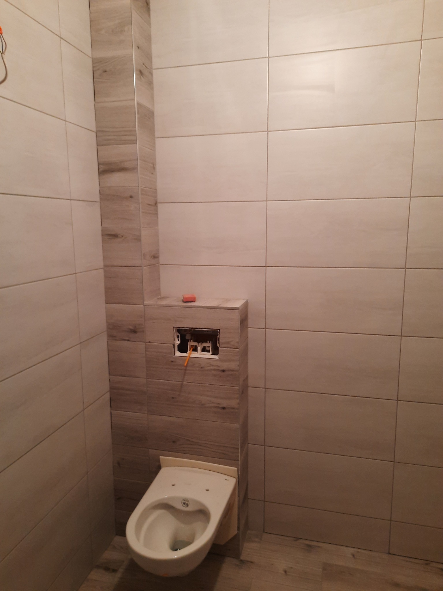 Niedokończona toaleta w łazience z jasnymi, drewnopodobnymi płytkami, widoczne przewody i puszka instalacyjna za stelażem podtynkowym, na górze leży poziomica