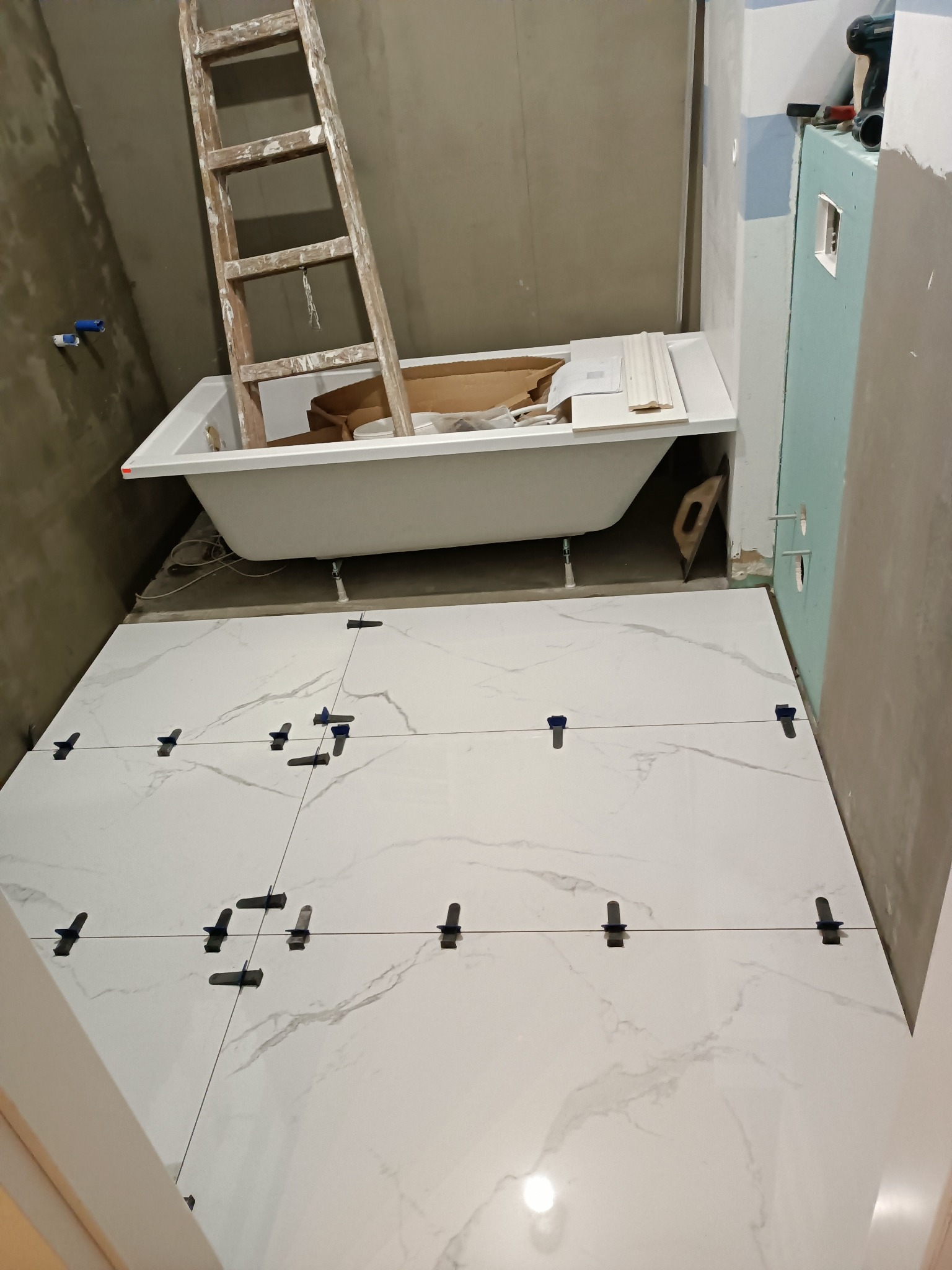Ułożone białe płytki podłogowe z imitacją marmuru w trakcie prac wykończeniowych łazienki, widoczne niebieskie klipsy poziomujące, drabina oparta o wannę, ściany w stanie surowym.