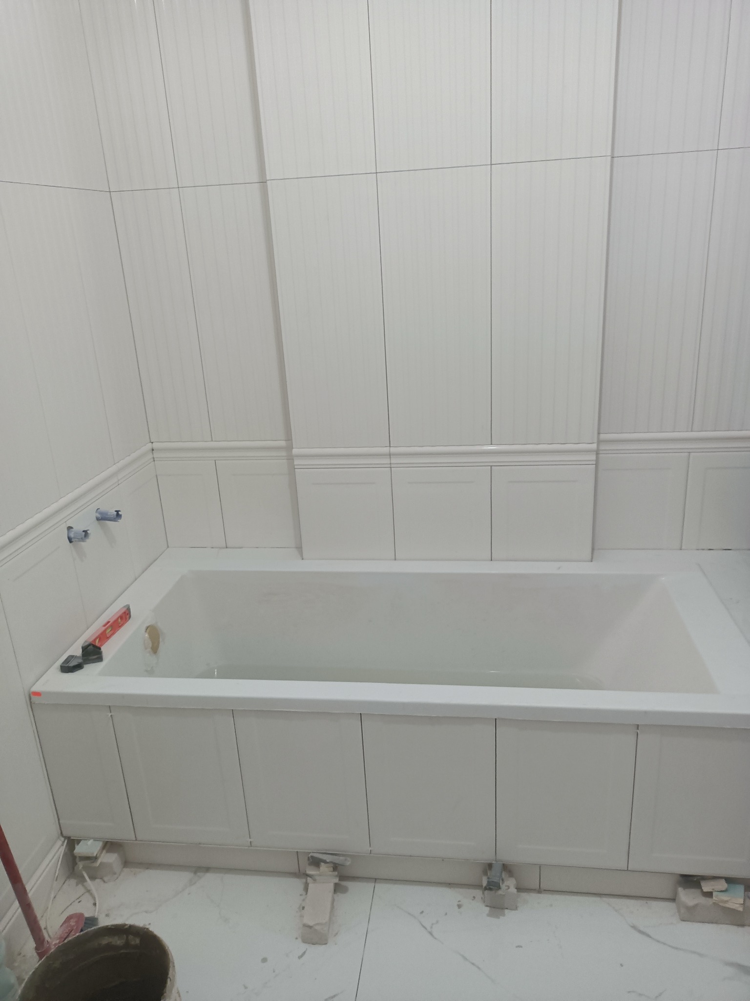 Biała wanna obudowana panelami, białe płytki ścienne z pionowymi rowkami, poziomica na wannie, wiadro z zaprawą w rogu, prace w toku.
