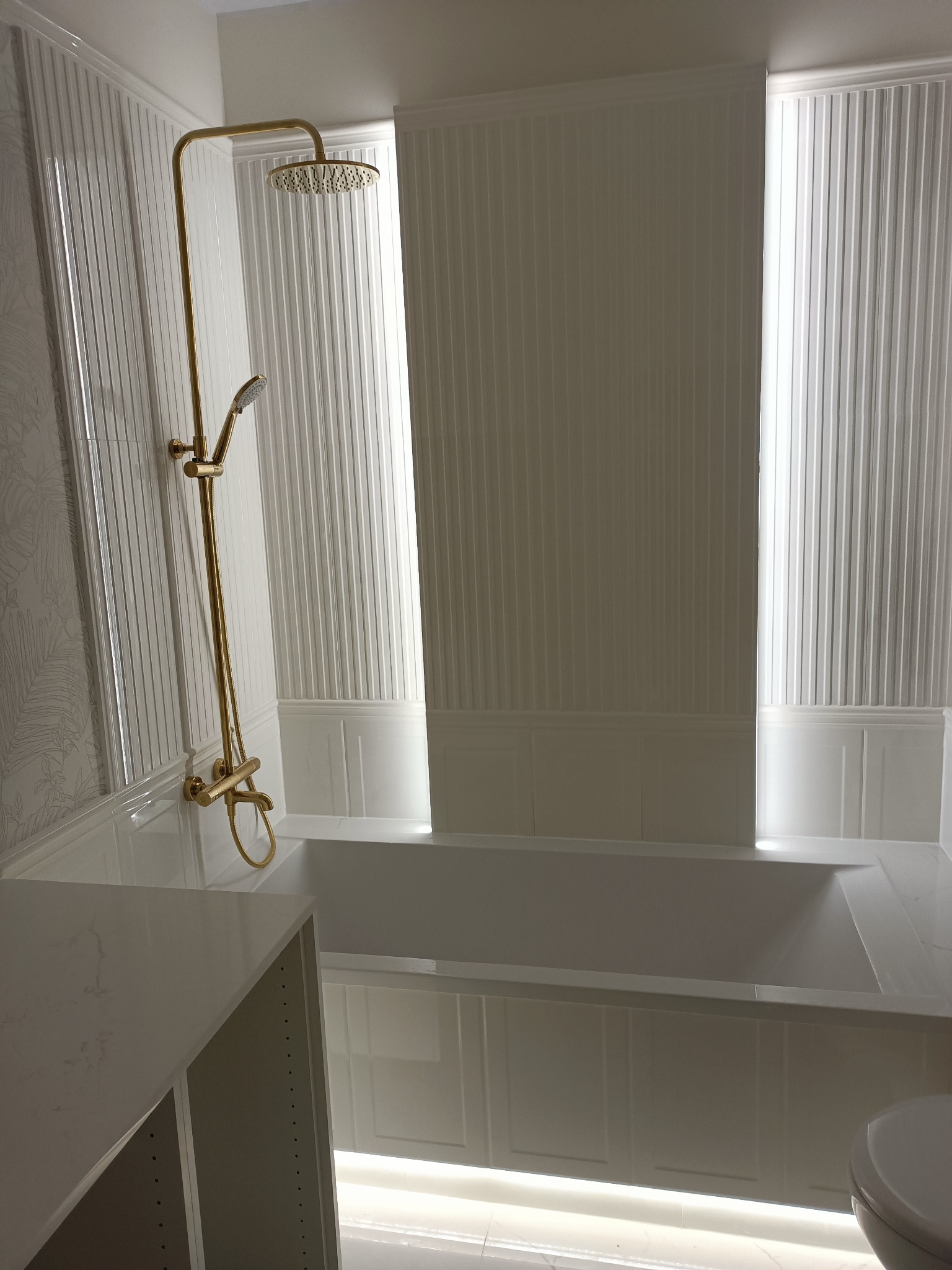 Elegancka, biała łazienka z wanną obudowaną panelami, złotą armaturą prysznicową i subtelnym oświetleniem LED przy podłodze i w niszach ściennych.