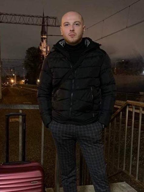 Mężczyzna w czarnej kurtce puchowej i spodniach w kratę, stojący na zewnątrz z walizką, w tle oświetlony budynek przypominający kościół i elementy infrastruktury kolejowej.