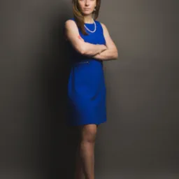 Portret kobiety w niebieskiej sukience i perłach, z założonymi rękami na szarym tle, sugerujący profesjonalizm i pewność siebie.