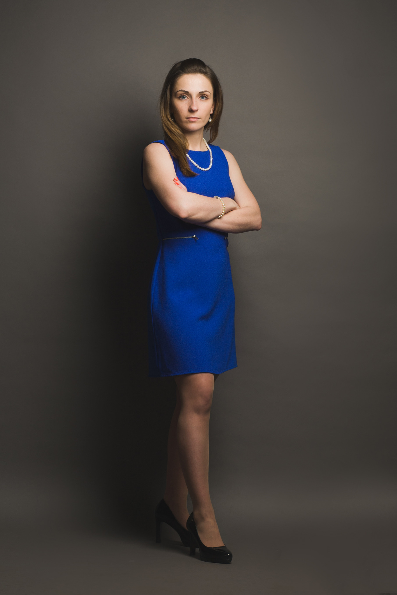 Portret kobiety w niebieskiej sukience i perłach, z założonymi rękami na szarym tle, sugerujący profesjonalizm i pewność siebie.
