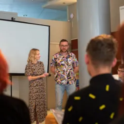 Prezenterzy podczas prezentacji na tle białego ekranu, widoczni słuchacze na pierwszym planie, mężczyzna w koszuli w geometryczne wzory i kobieta w kwiecistej sukience.