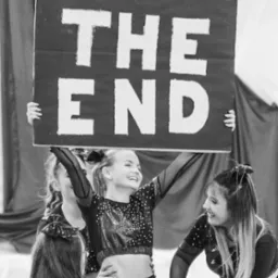 Czarno-białe zdjęcie grupy uśmiechniętych cheerleaderek trzymających nad głowami duży karton z napisem 'THE END' podczas występu.