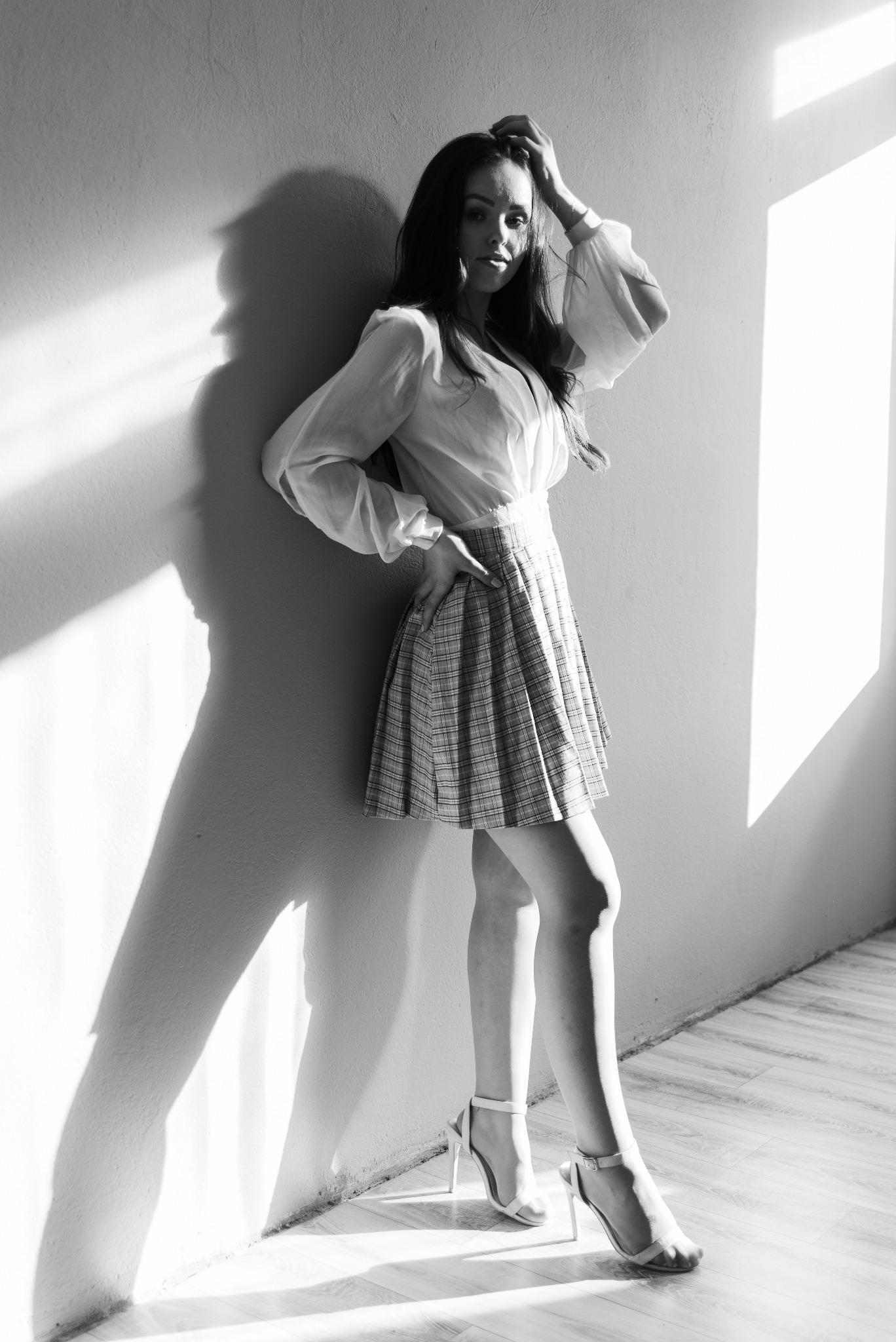 Czarno-białe zdjęcie kobiety w bluzce z bufiastymi rękawami i spódnicy w kratę, opierającej się o ścianę w silnym świetle, z wyraźnym cieniem na ścianie.