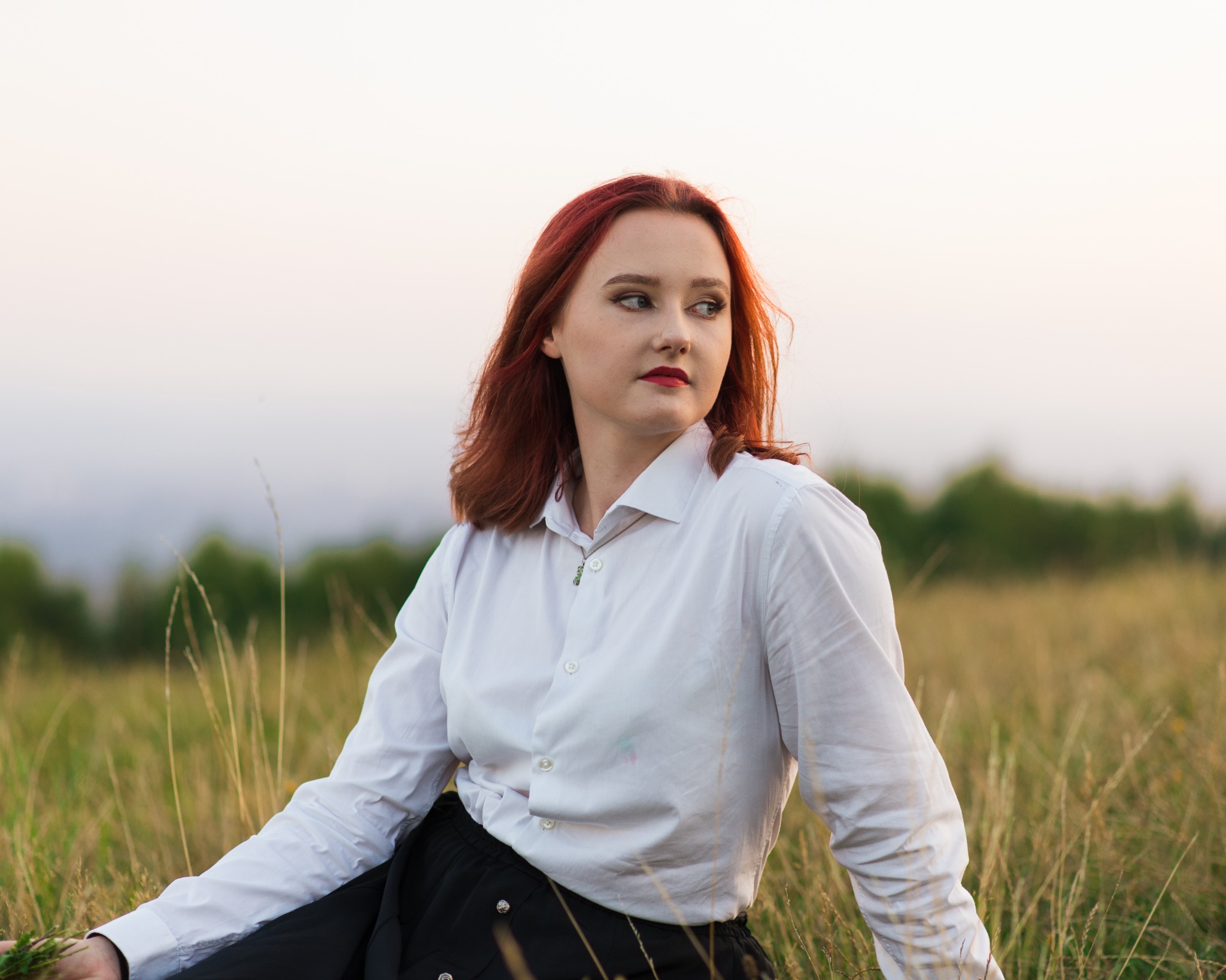 Portret kobiety o rudych włosach i czerwonych ustach w białej koszuli, siedzącej na łące w promieniach zachodzącego słońca.