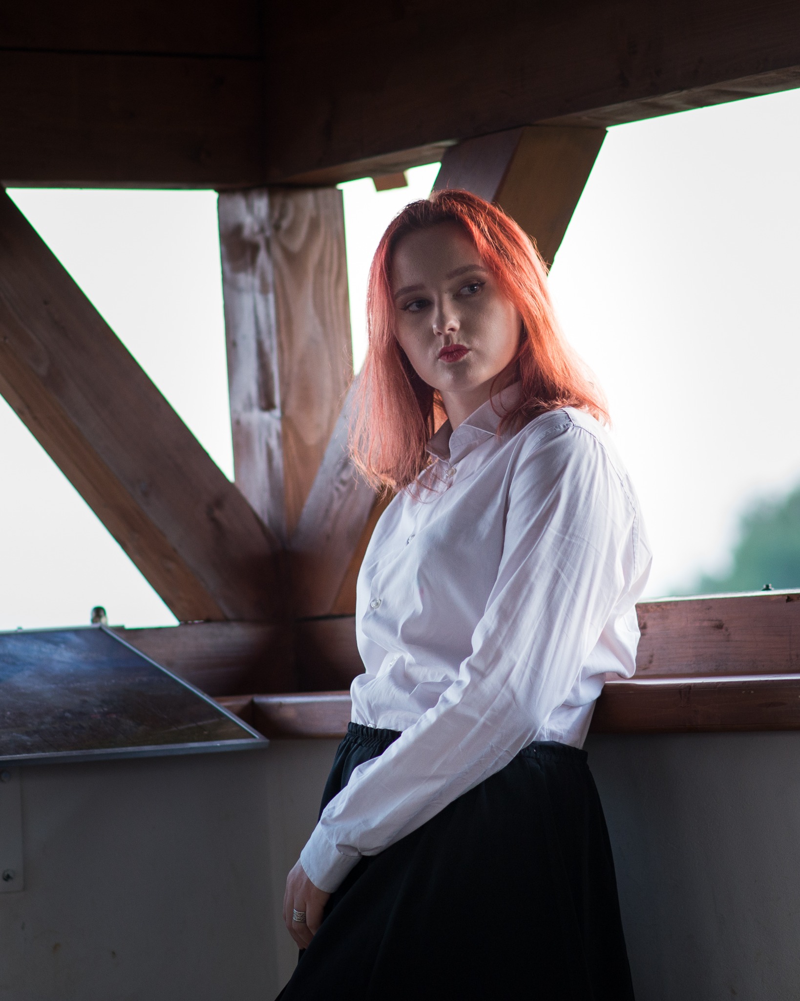 Portret kobiety z rudymi włosami w białej koszuli i czarnej spódnicy, opierającej się o drewnianą konstrukcję w plenerze.