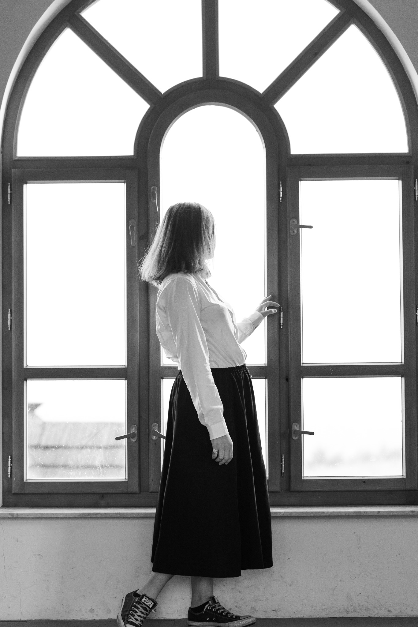 Czarno-białe zdjęcie kobiety stojącej przy oknie z łukowym szprosem, ubrana w białą koszulę, długą czarną spódnicę i trampki, odwrócona tyłem do aparatu, z ręką na klamce okna.