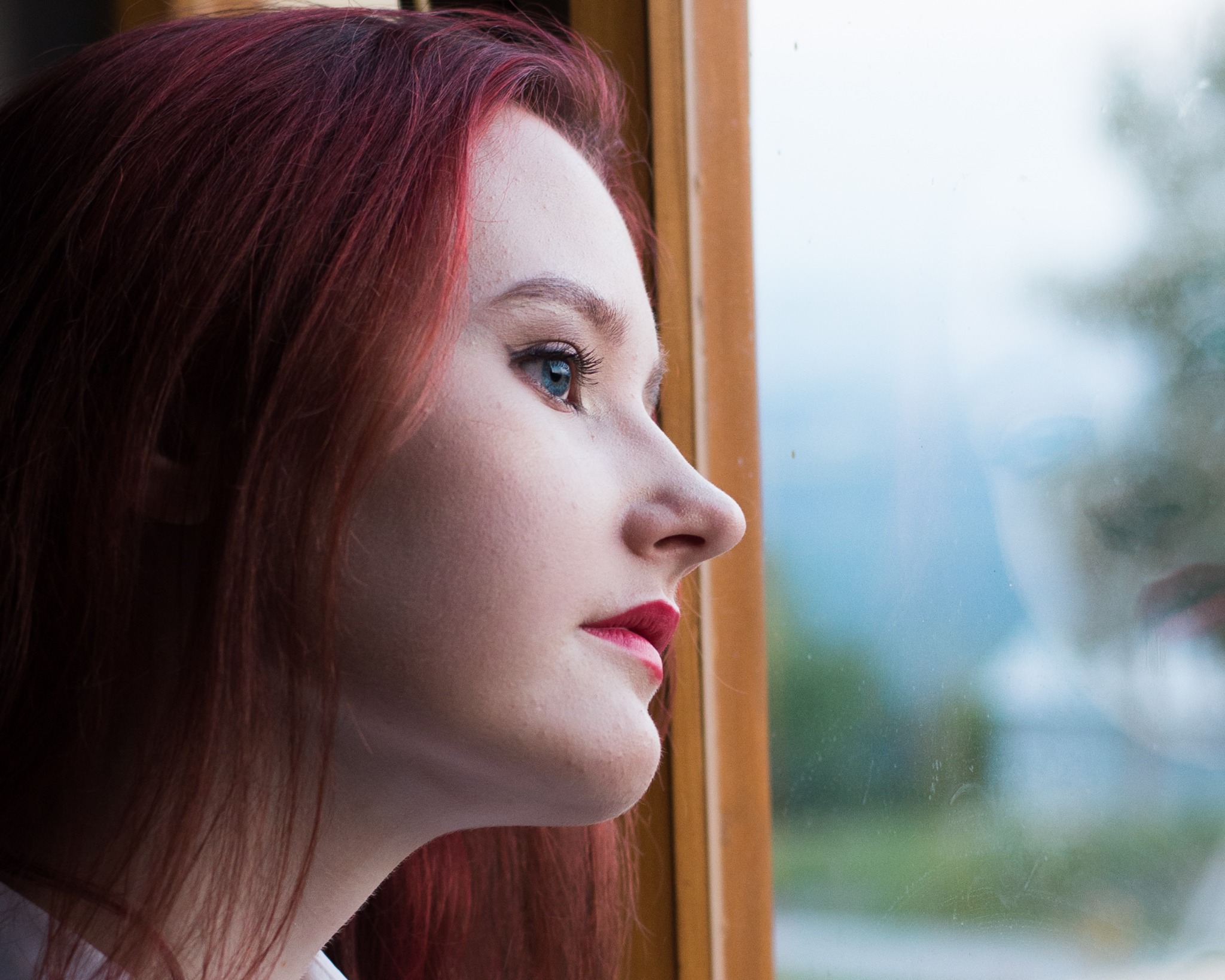 Portret z profilu młodej kobiety z rudymi włosami i niebieskimi oczami, spoglądającej przez okno z drewnianą ramą na rozmyty krajobraz.