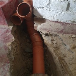 FMBUD - Pomarańczowe rury kanalizacyjne ułożone w wykopie przy fundamencie z cegły, widok z góry, surowy stan.
