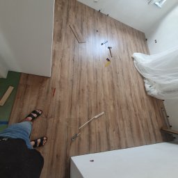 FMBUD - Montaż paneli podłogowych z wzorem drewna w pokoju ze skosami, widoczne narzędzia, elementy montażowe, fragment podłogi z innym wykończeniem i osoba w sandałach.