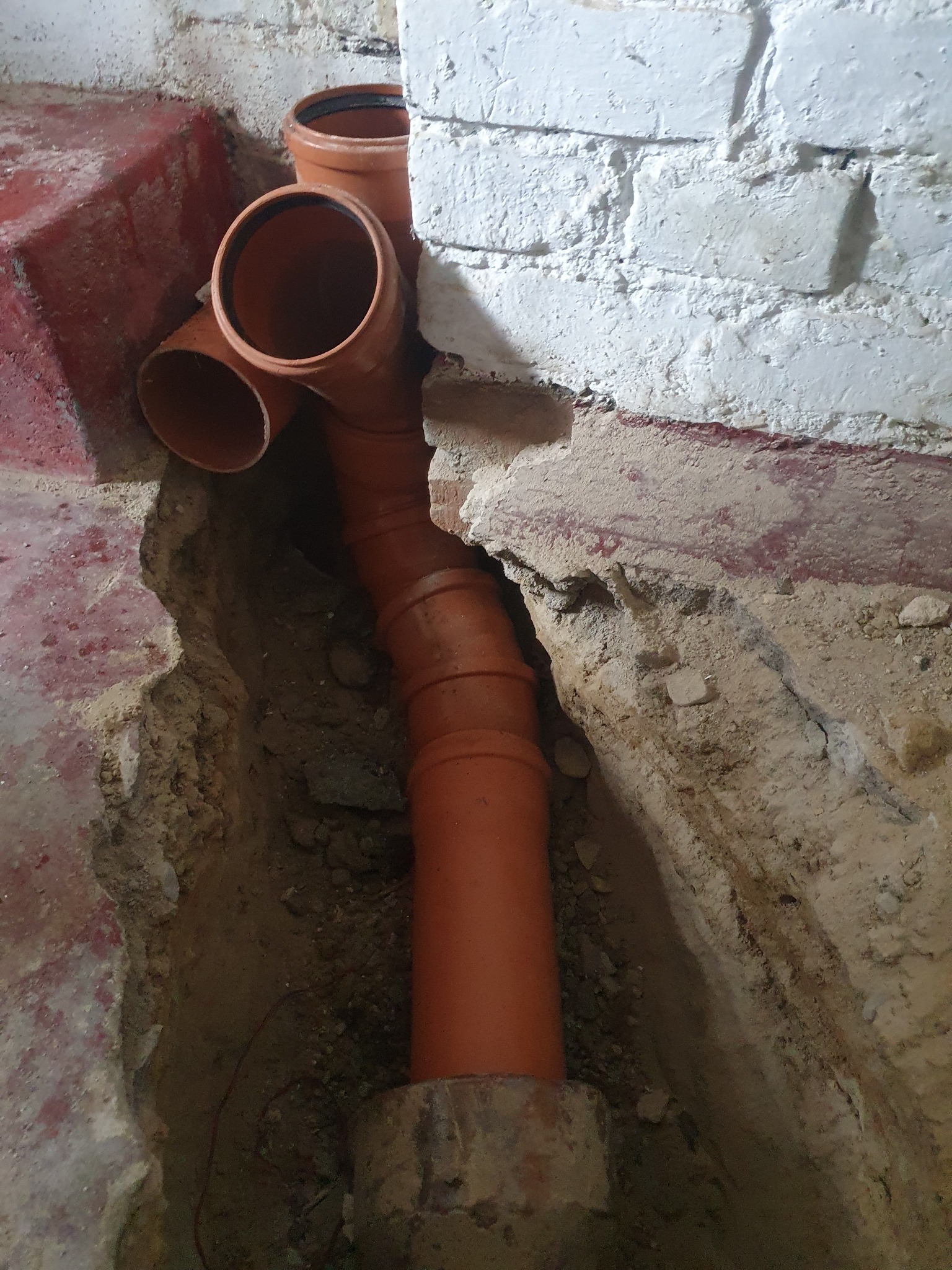 Pomarańczowe rury kanalizacyjne ułożone w wykopie przy fundamencie z cegły, widok z góry, surowy stan.