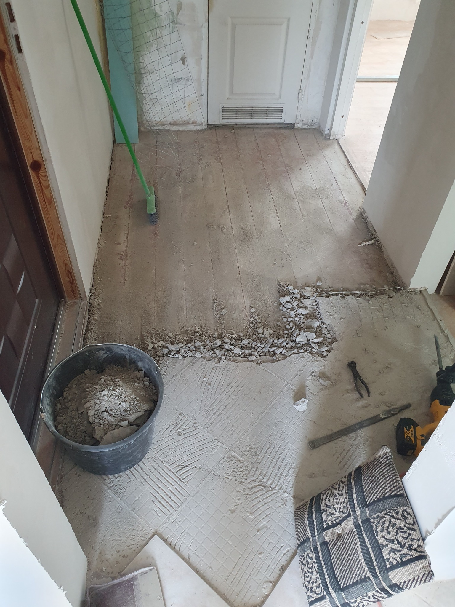 Demontaż starej podłogi w korytarzu: gruz, wiadro z resztkami, narzędzia i poduszka w rogu podczas prac remontowych.