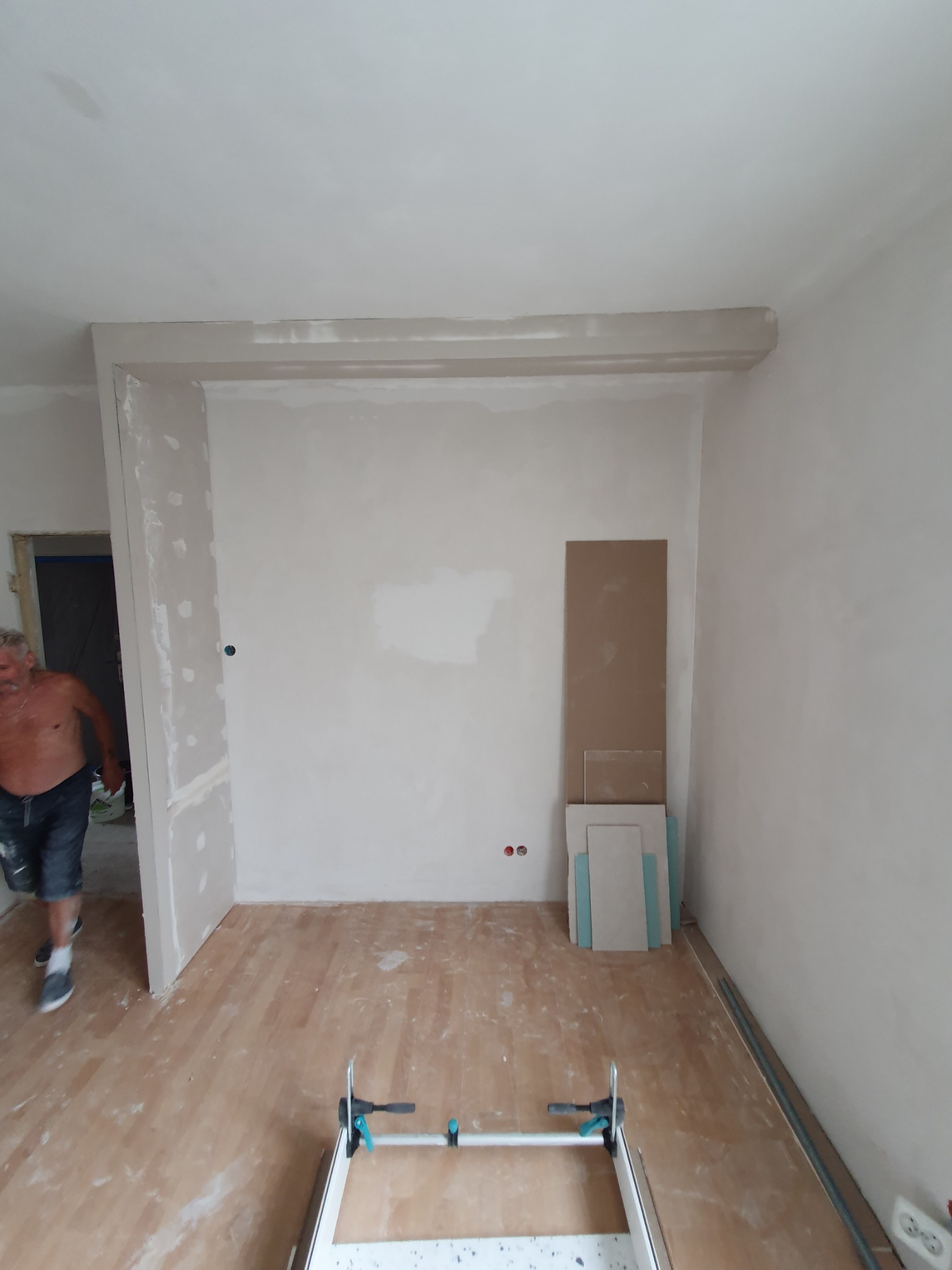 Wnętrze w trakcie prac wykończeniowych: ściany po szpachlowaniu, widoczne płyty karton-gips oparte o ścianę, podłoga drewniana częściowo zakryta pyłem, pracownik w tle.