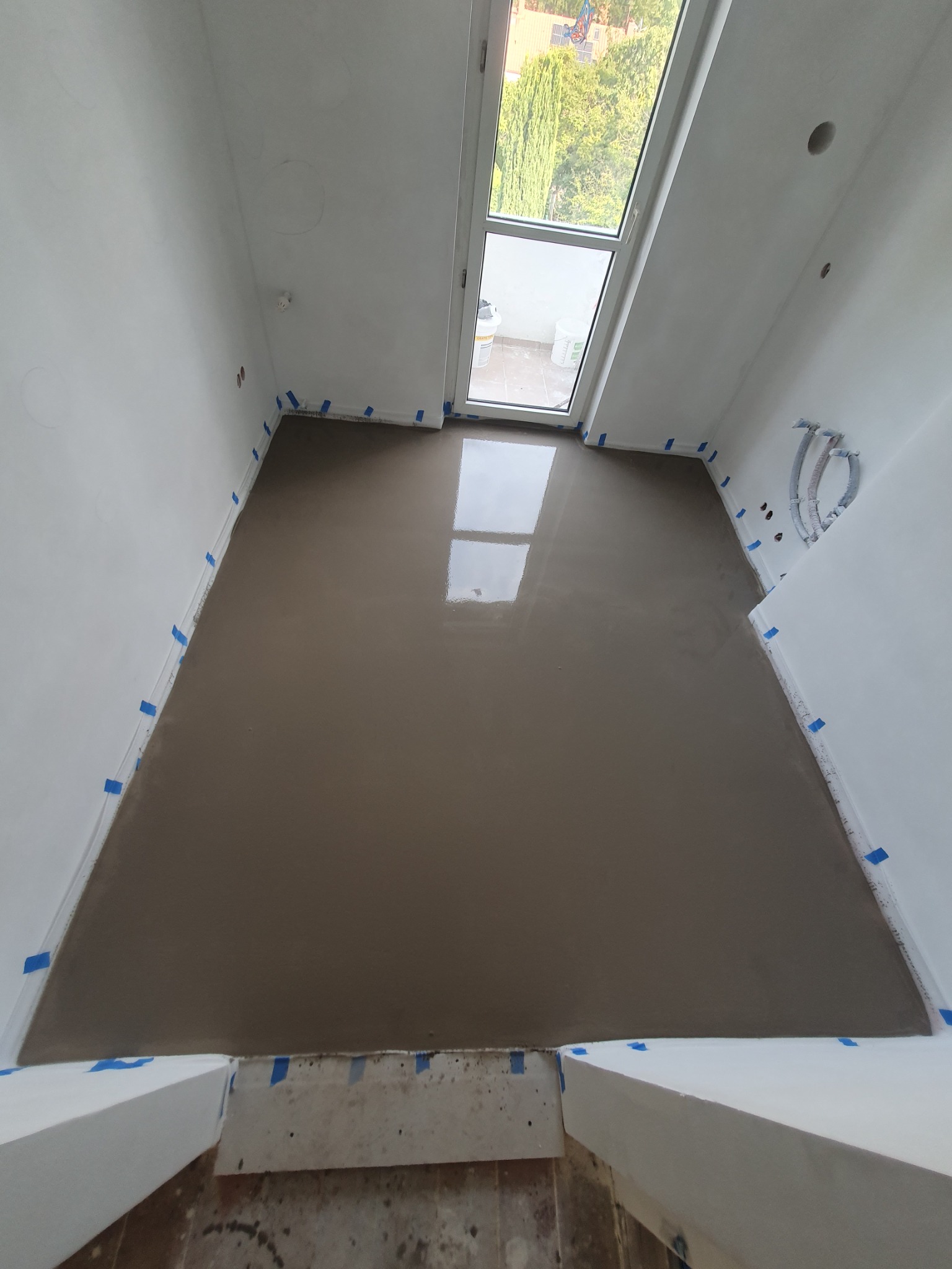 Wylany, mokry, samopoziomujący podkład podłogowy w pomieszczeniu z oknem dachowym, zabezpieczonym taśmą malarską przy ścianach.