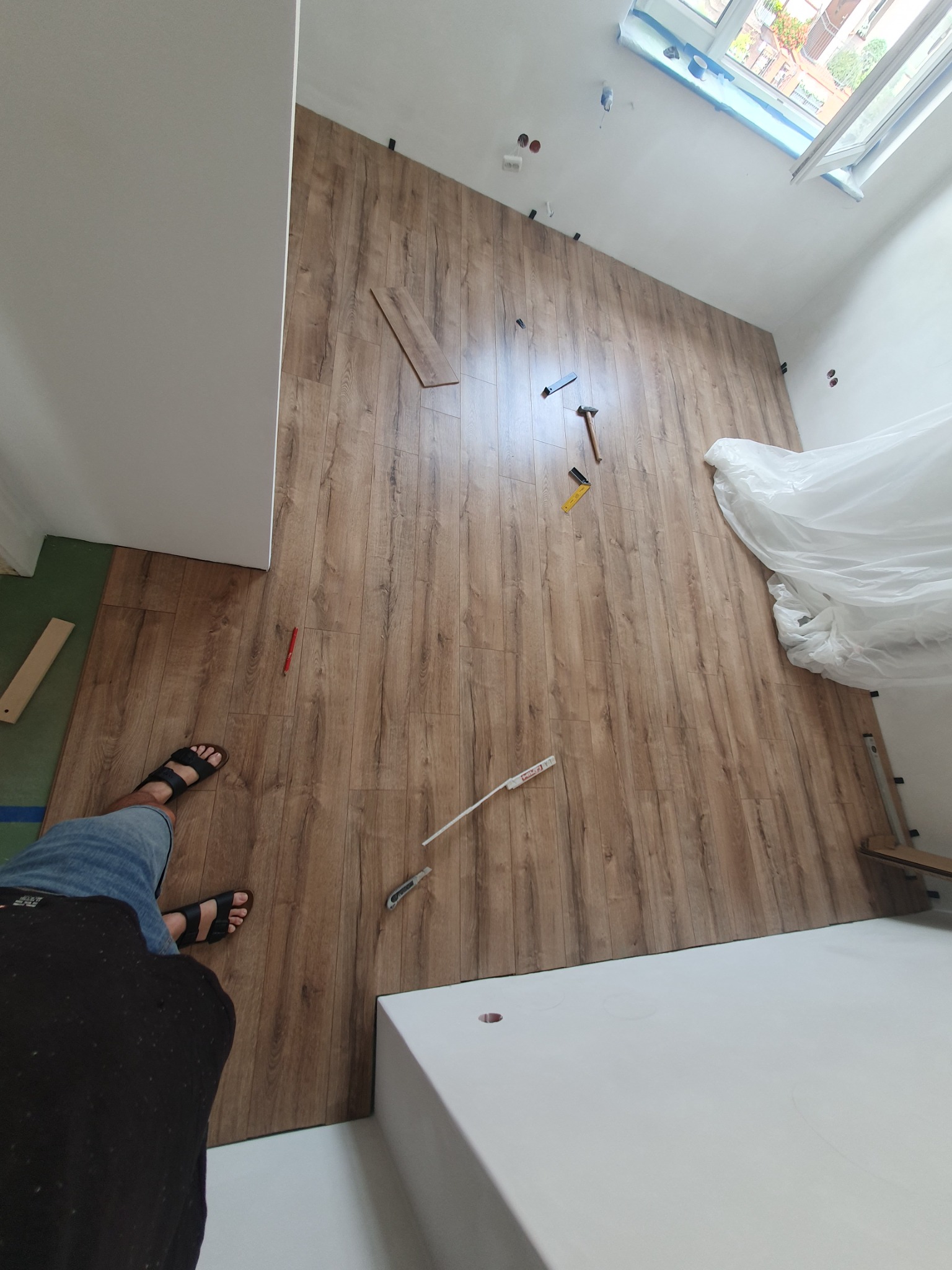 Montaż paneli podłogowych z wzorem drewna w pokoju ze skosami, widoczne narzędzia, elementy montażowe, fragment podłogi z innym wykończeniem i osoba w sandałach.