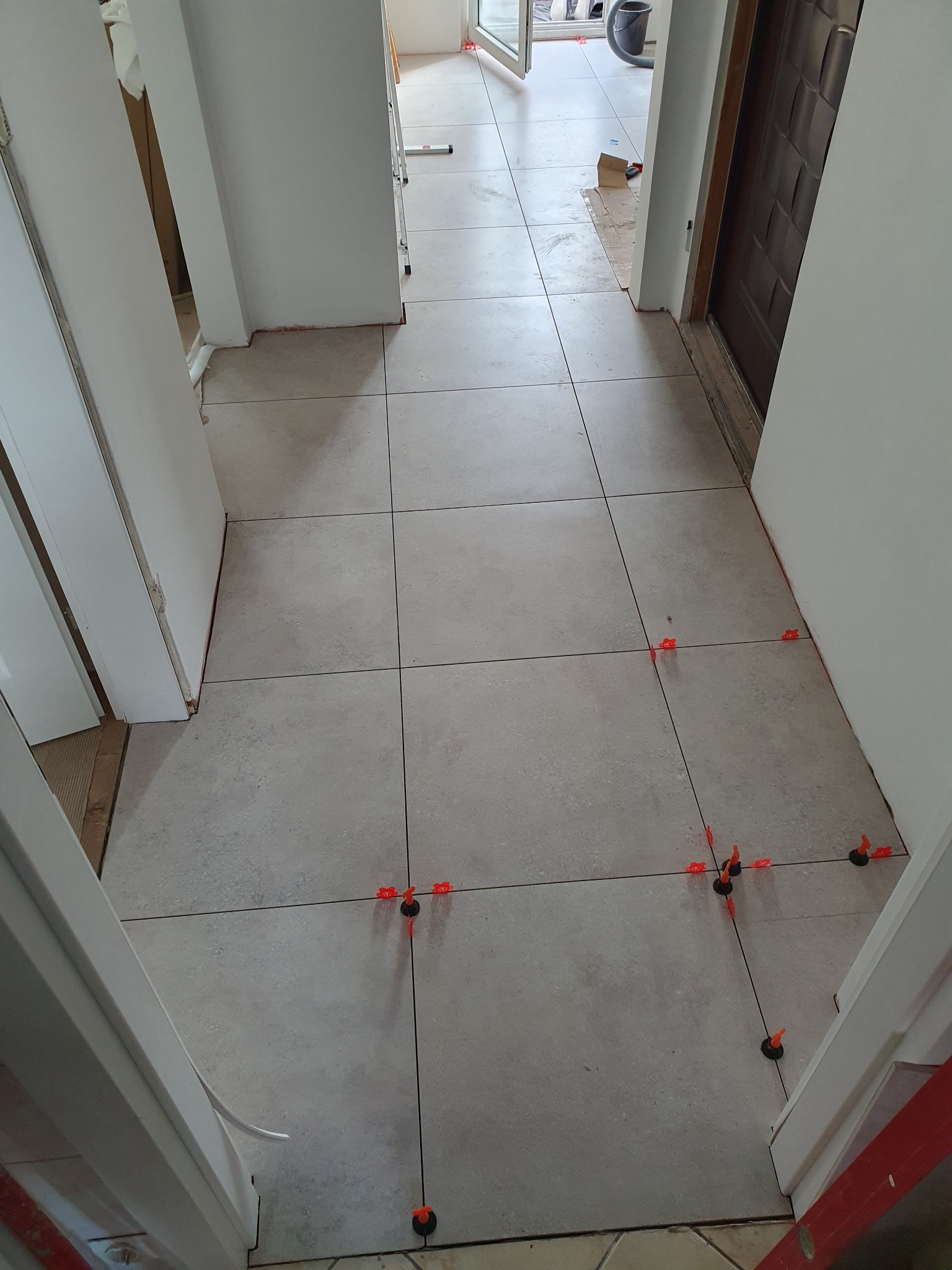 Układanie szarych płytek podłogowych w korytarzu, z użyciem pomarańczowych klinów poziomujących, widok z góry na postęp prac.