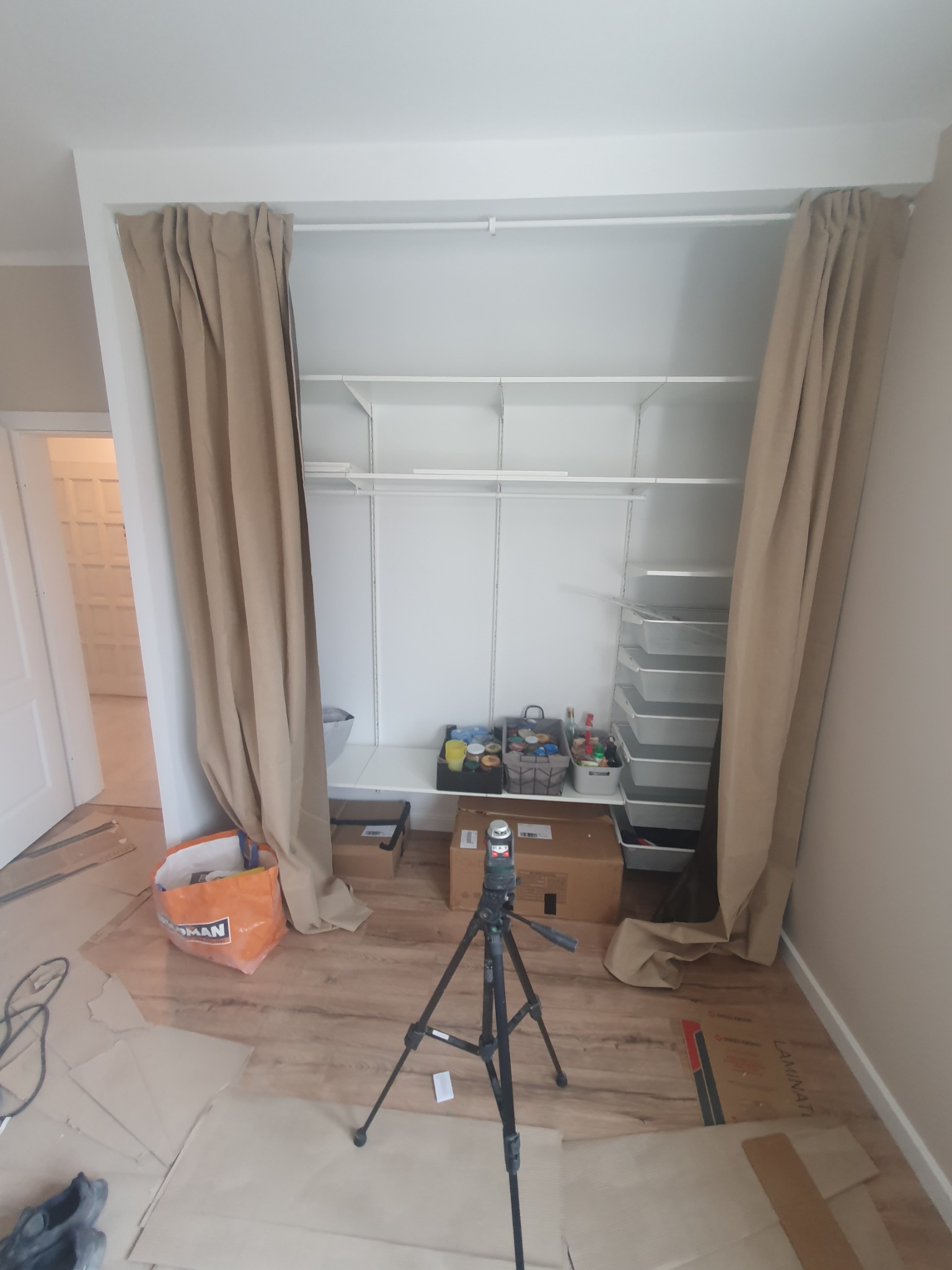 Montaż metalowych półek wnękowych z zasłoniętymi beżowymi zasłonami, widoczny laser krzyżowy na statywie i zabezpieczona podłoga.