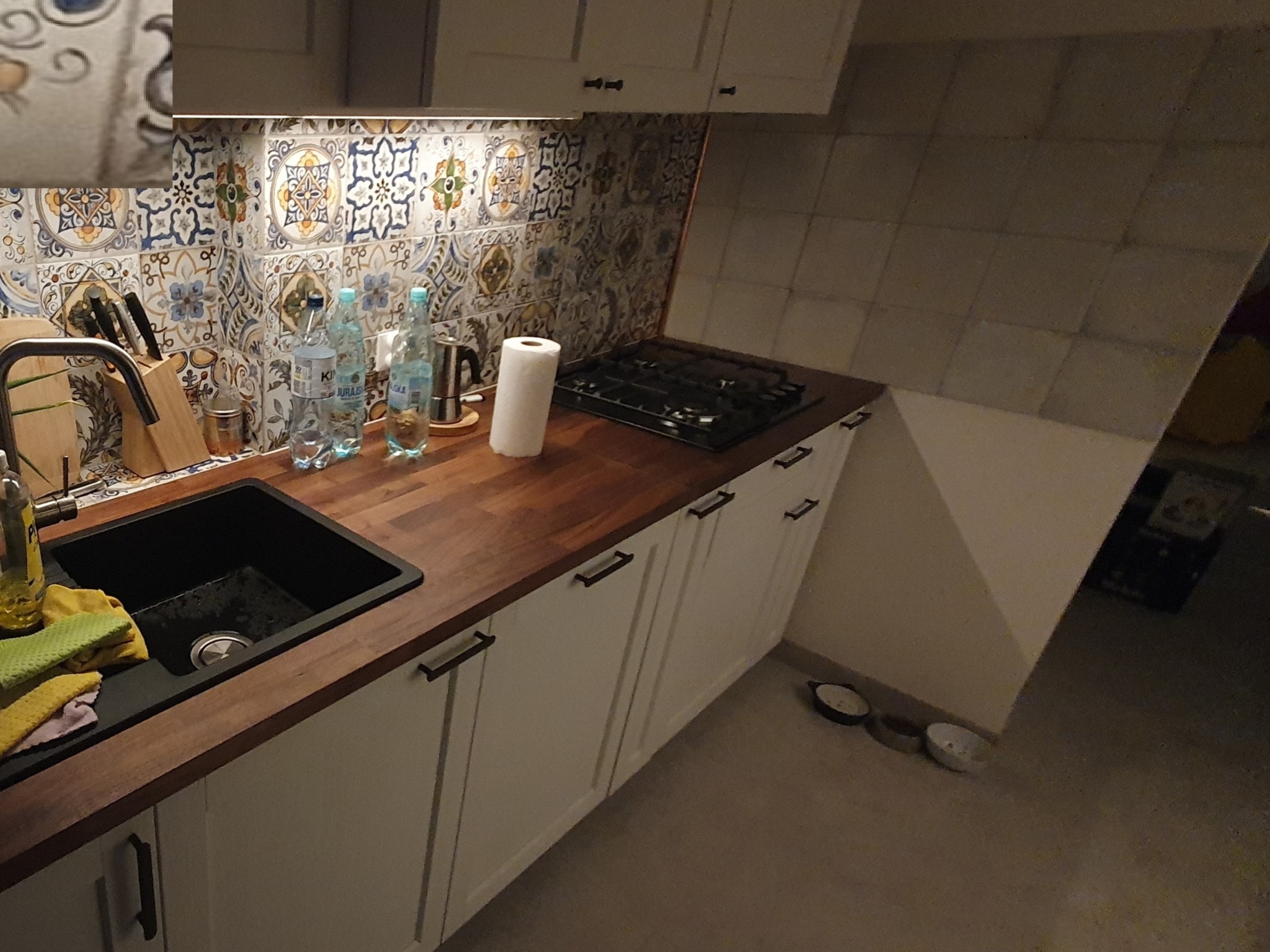 Nowoczesna kuchnia z ciemnym blatem, białymi szafkami z czarnymi uchwytami, kolorową mozaiką nad blatem, czarnym zlewem i płytą gazową.