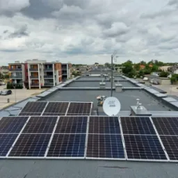 Panele słoneczne na płaskim dachu budynku wielorodzinnego, widok z góry na tle zabudowy miejskiej i pochmurnego nieba.