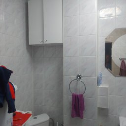 M-Bud Marek Kujawa - Biała łazienka po remoncie: płytki imitujące marmur, szafka wisząca, okrągły wieszak na ręcznik. W lustrze odbicie osoby. Widoczny fragment toalety i akcesoriów.