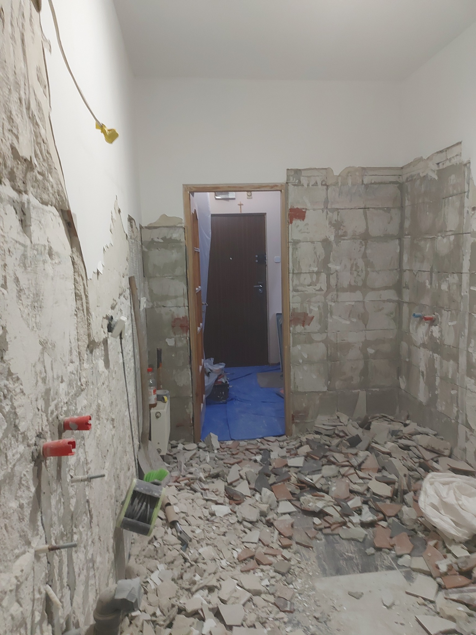 Łazienka w trakcie remontu: zerwane płytki, odsłonięte instalacje, gruz na podłodze. Widoczne narzędzia i fragmenty instalacji hydraulicznej.
