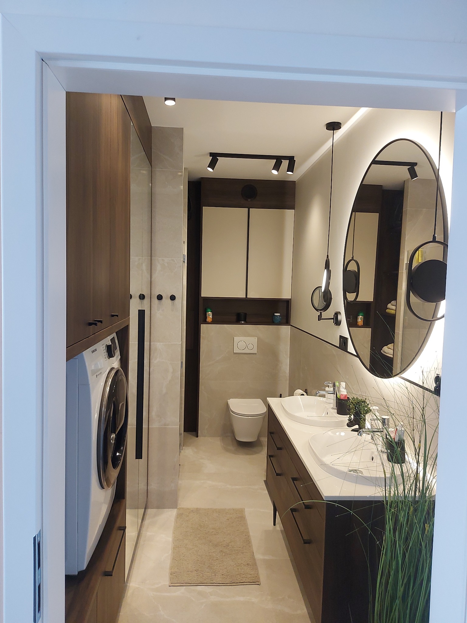 Nowoczesna łazienka z pralką w zabudowie, podwójną umywalką, owalnym lustrem z podświetleniem LED i minimalistycznym oświetleniem punktowym.