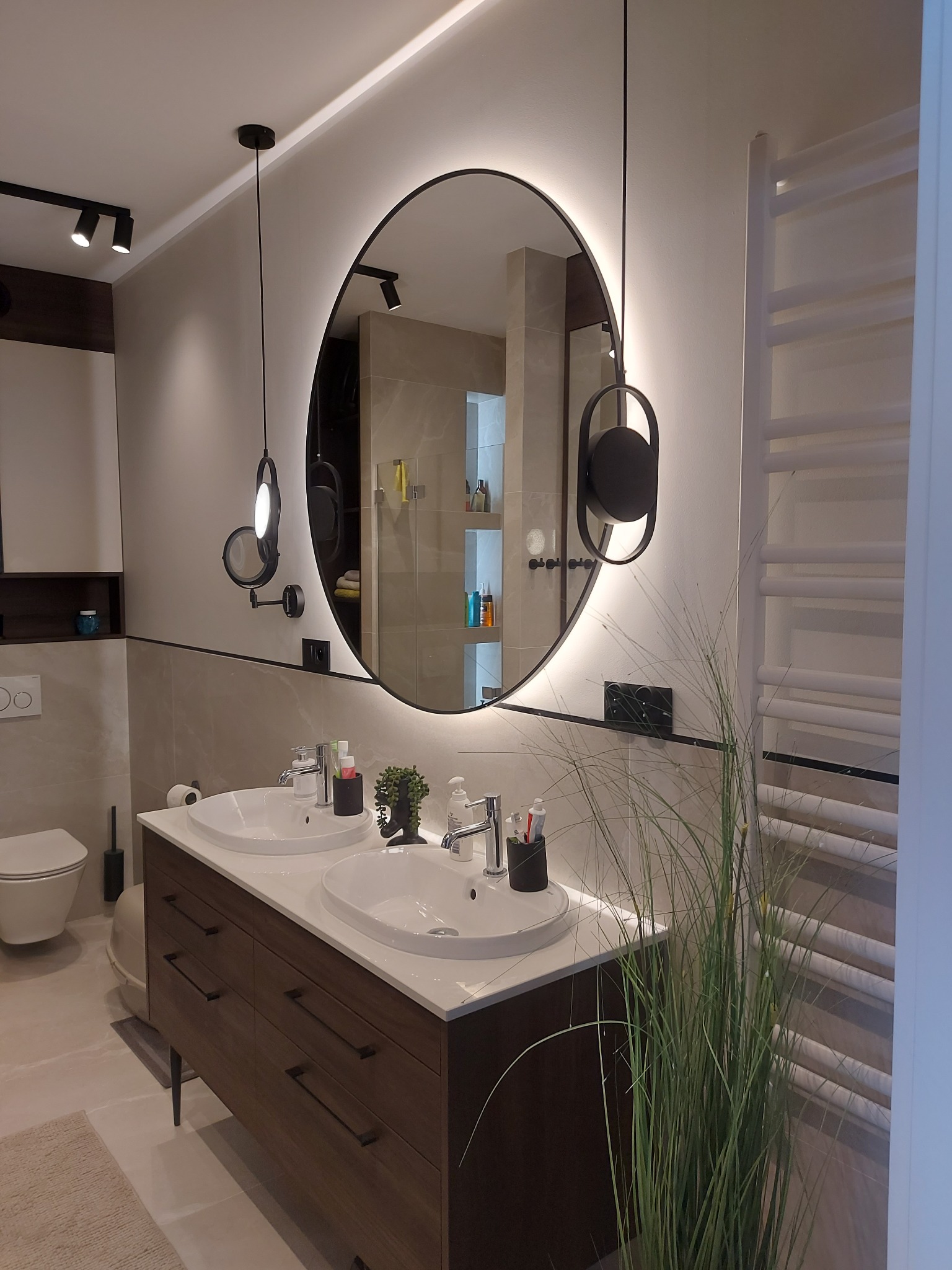 Nowoczesna łazienka z okrągłym lustrem podświetlanym LED, umywalką nablatową i grzejnikiem drabinkowym. Minimalistyczny design, ciepłe oświetlenie.