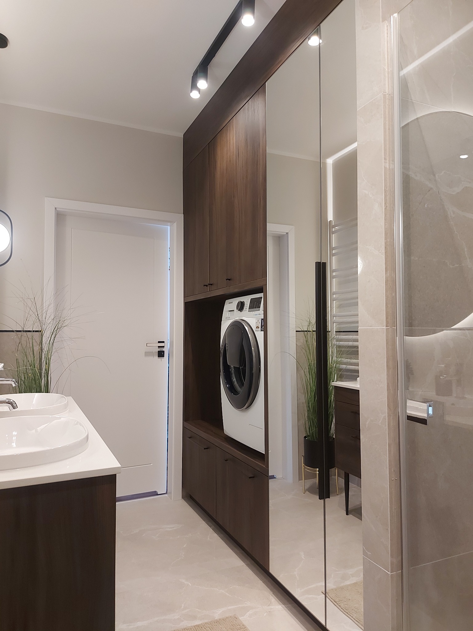 Nowoczesna łazienka z zabudową z ciemnego drewna, w której umieszczono pralkę. Umywalka z białym blatem i duże lustro optycznie powiększają przestrzeń.