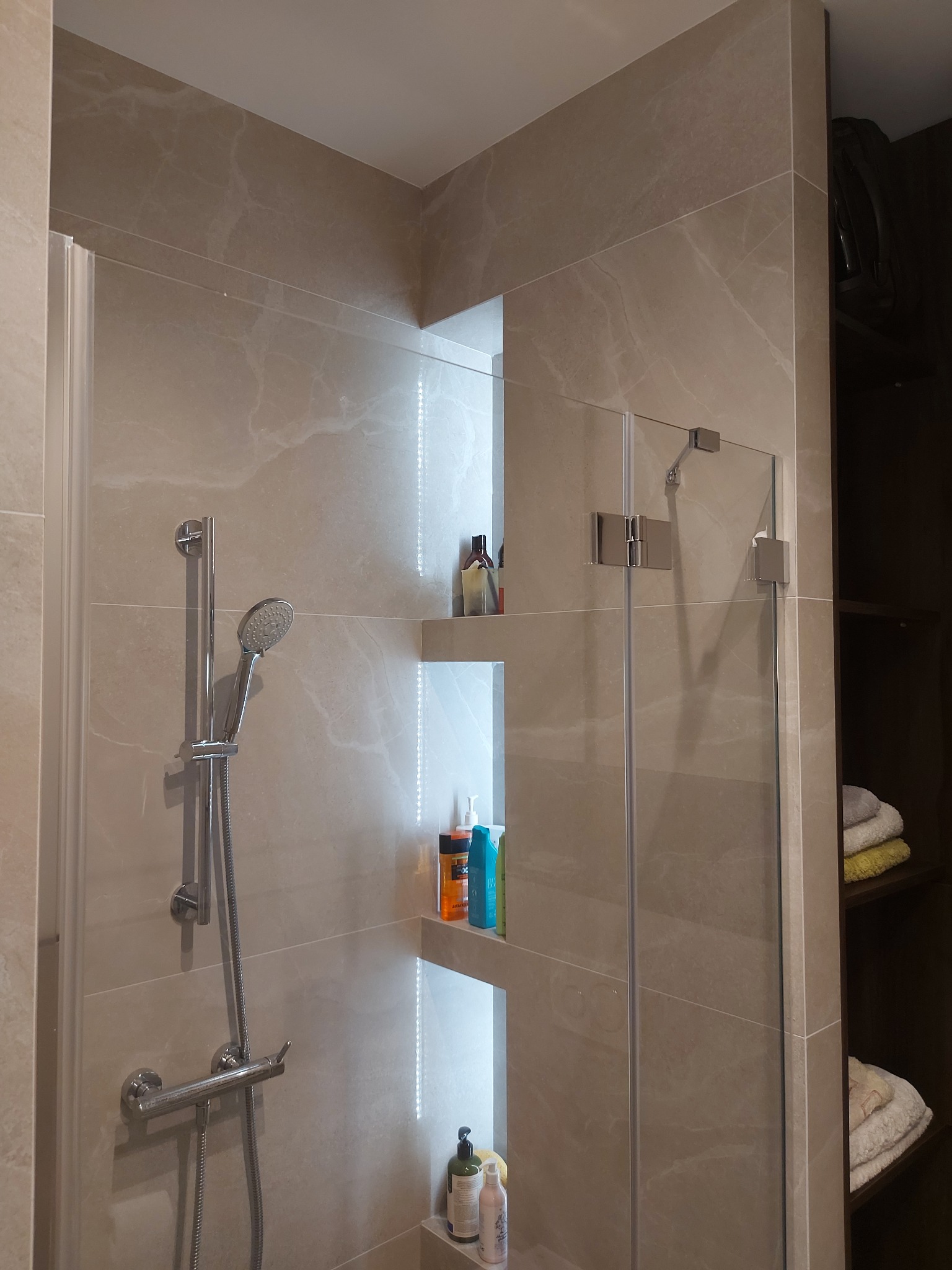 Beżowa łazienka z kabiną prysznicową, półkami z oświetleniem LED i armaturą w kolorze chromu. Widoczne kosmetyki i ręczniki.