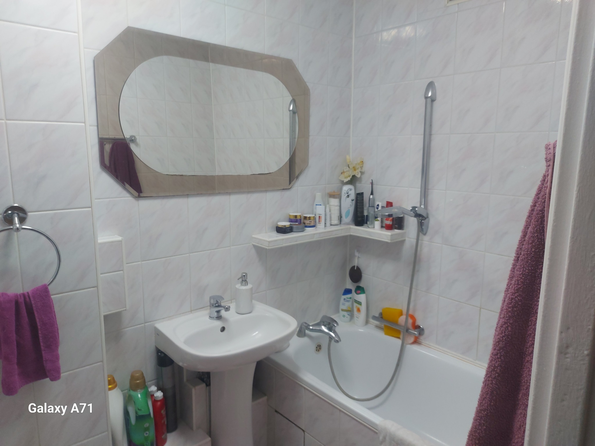 Wnętrze łazienki z białą armaturą i płytkami. Umywalka na postumencie, wanna z prysznicem, lustro o nietypowym kształcie. Na półce kosmetyki i dekoracje.