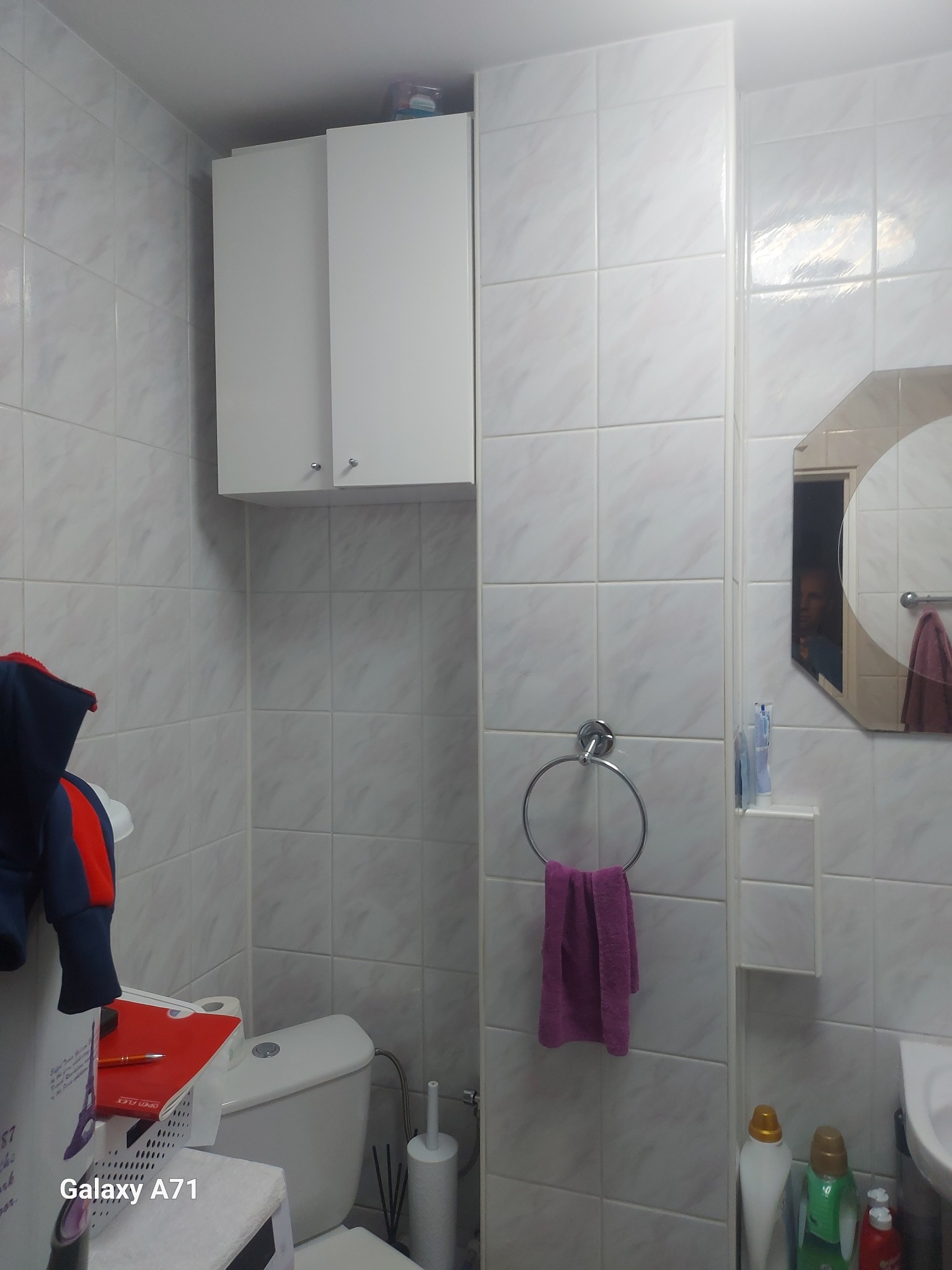 Biała łazienka po remoncie: płytki imitujące marmur, szafka wisząca, okrągły wieszak na ręcznik. W lustrze odbicie osoby. Widoczny fragment toalety i akcesoriów.