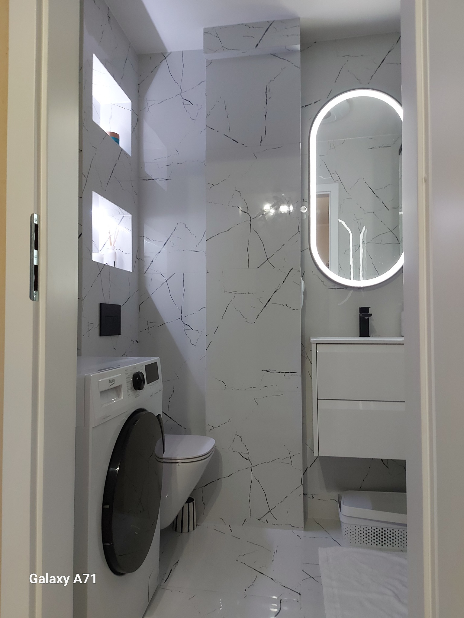 Nowoczesna łazienka z białą armaturą, marmurowymi płytkami i podświetlanym lustrem. Widoczna pralka i wnęki z dekoracjami. Minimalistyczny design.