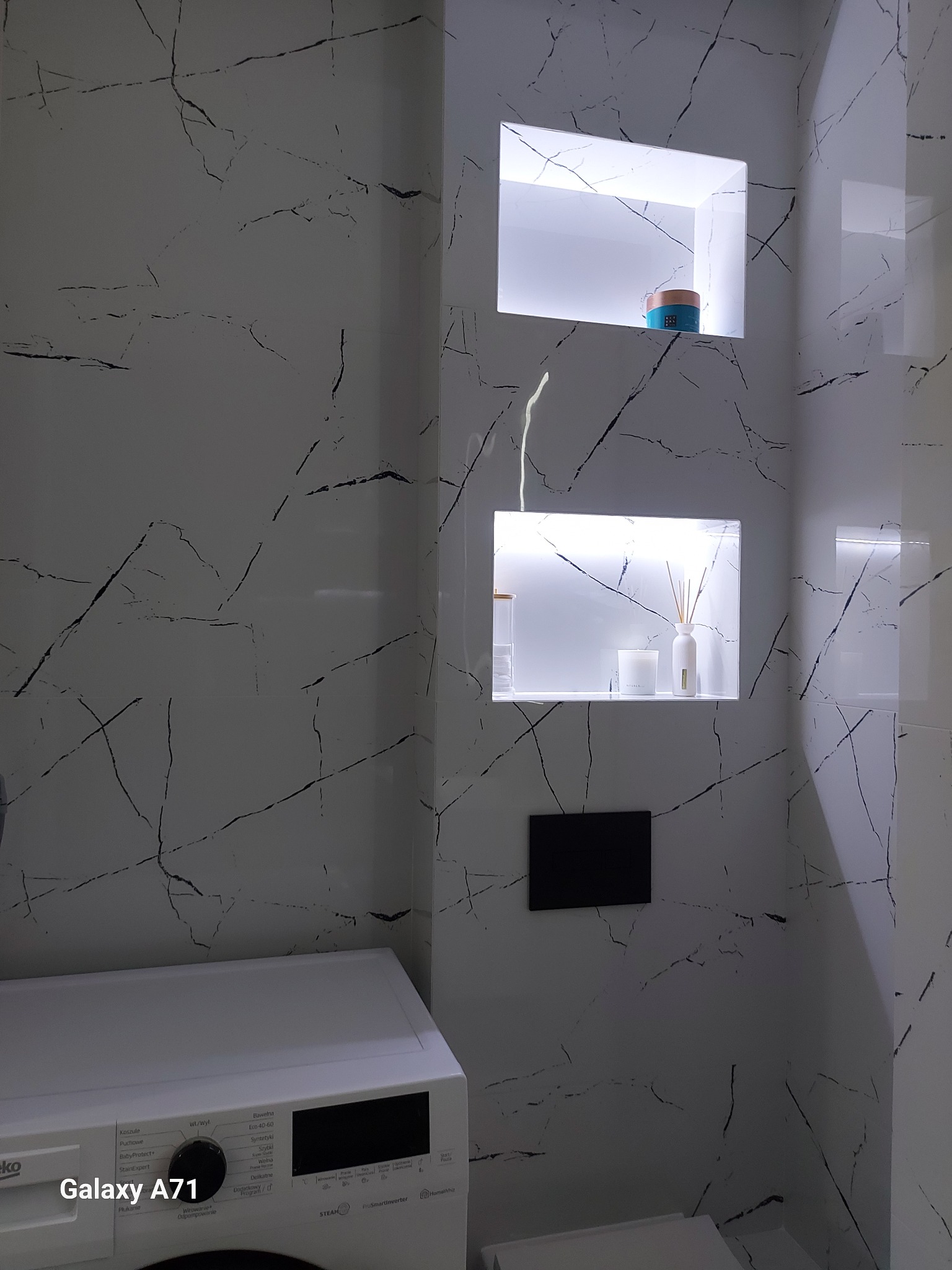 Biała łazienka z płytkami imitującymi marmur, podświetlane wnęki z dekoracjami, pralka Beko Galaxy A71. Minimalistyczny design.