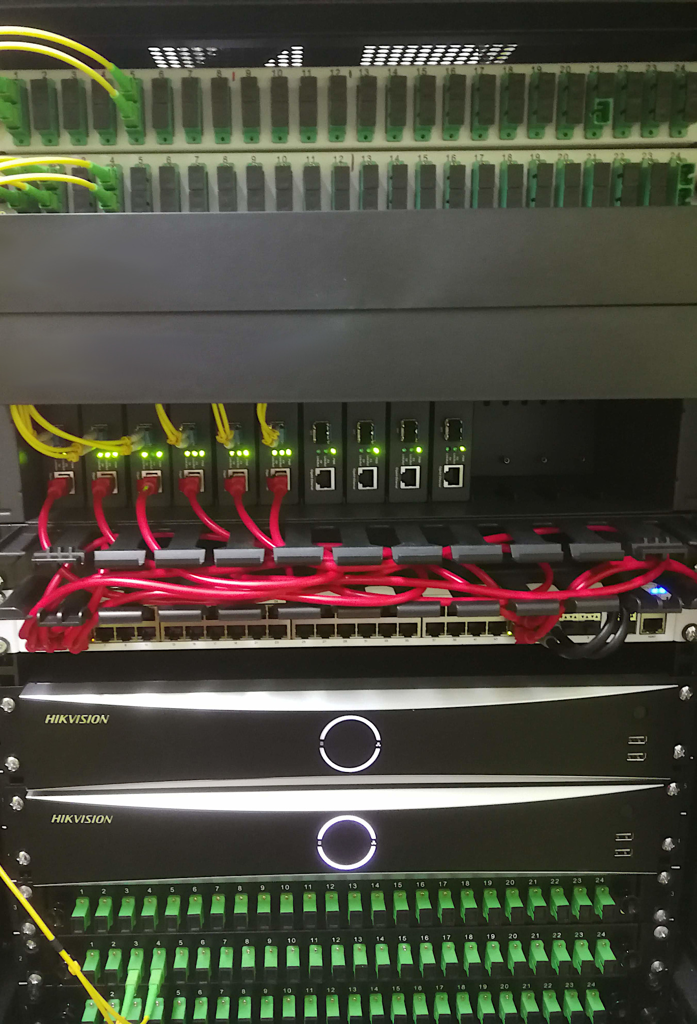 Szafa serwerowa z uporządkowanymi kablami światłowodowymi (zielone i żółte) oraz patchcordami (czerwone), widoczne urządzenia Hikvision z podświetlanymi okręgami, oświetlenie LED sygnalizujące...