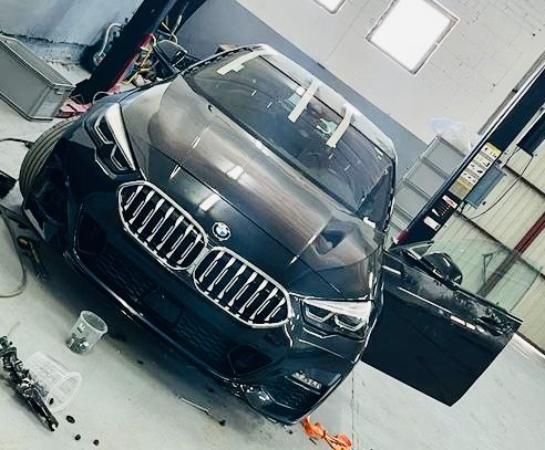 Ciemny samochód BMW z otwartymi drzwiami w warsztacie, zabezpieczony taśmą malarską na szybie, z widocznymi narzędziami lakierniczymi i kubkami na podłodze.