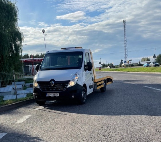 Biała laweta Renault Master z żółtymi elementami platformy, zaparkowana na asfalcie w pobliżu stacji benzynowej i drogi, w tle widoczna wieża telekomunikacyjna pod błękitnym niebem z chmurami.