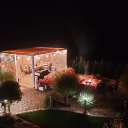 Oświetlona altana ogrodowa z grillem i stołem, w tle palenisko z płomieniami, nocne oświetlenie ścieżki, dekoracyjne drzewa i krzewy.