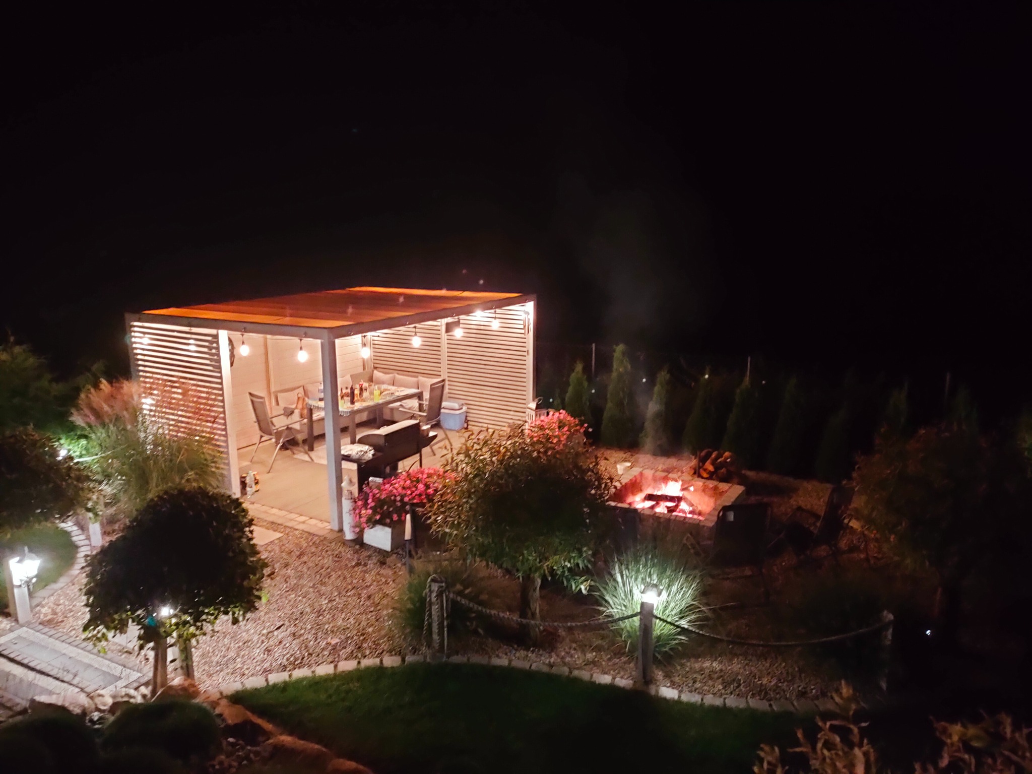 Oświetlona altana ogrodowa z grillem i stołem, w tle palenisko z płomieniami, nocne oświetlenie ścieżki, dekoracyjne drzewa i krzewy.