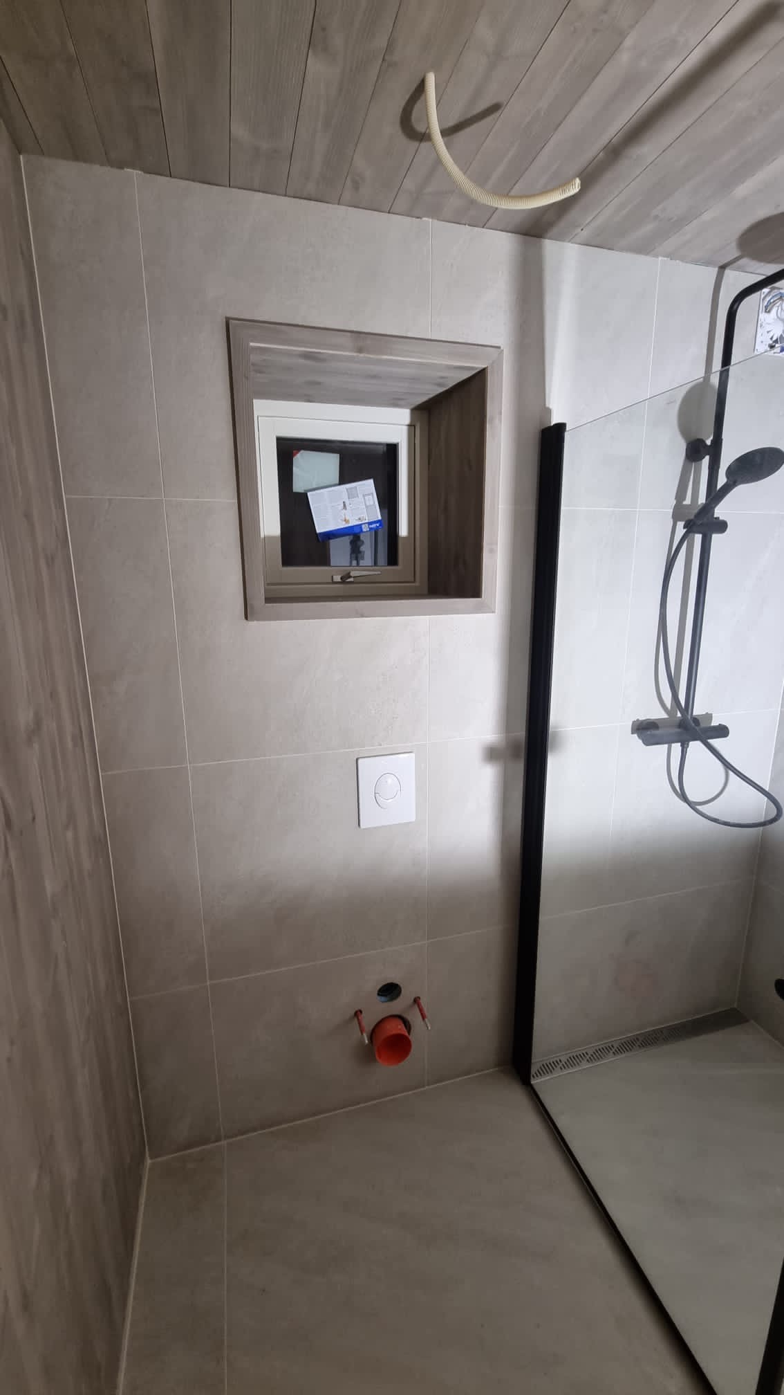 Szare płytki w łazience z zamontowanym stelażem podtynkowym WC w zabudowie, widoczny syfon i rury, oraz kabina prysznicowa z czarnymi profilami.