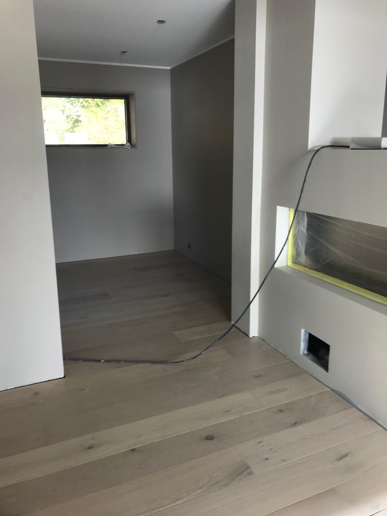 Wnętrze w trakcie wykańczania z jasną drewnianą podłogą, świeżo pomalowanymi ścianami w odcieniach szarości i białymi listwami przypodłogowymi, zabezpieczonym otworem w ścianie taśmą malarską...