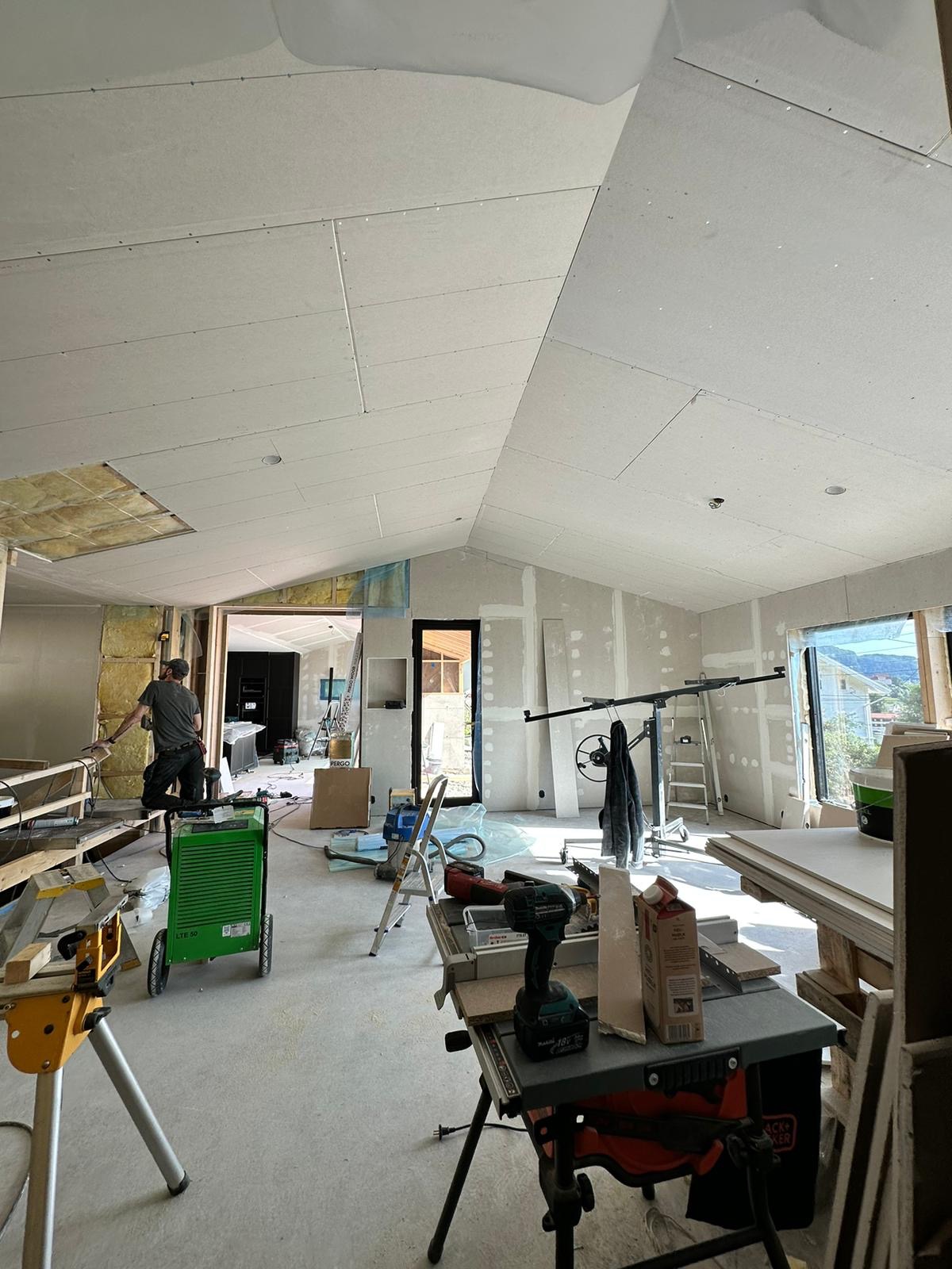 Wnętrze w trakcie remontu: montaż płyt karton-gips na skośnym suficie, widoczne narzędzia, drabina, podnośnik do płyt, oraz fragment ocieplenia wełną mineralną w ścianie.