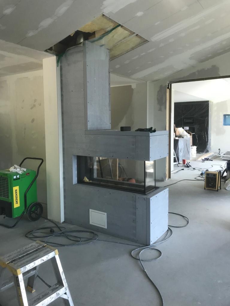 Wnętrze w trakcie wykończenia z widocznym kominkiem obudowanym płytami, otwór w suficie z widocznym ociepleniem, zielony osuszacz powietrza i drabina w pomieszczeniu.