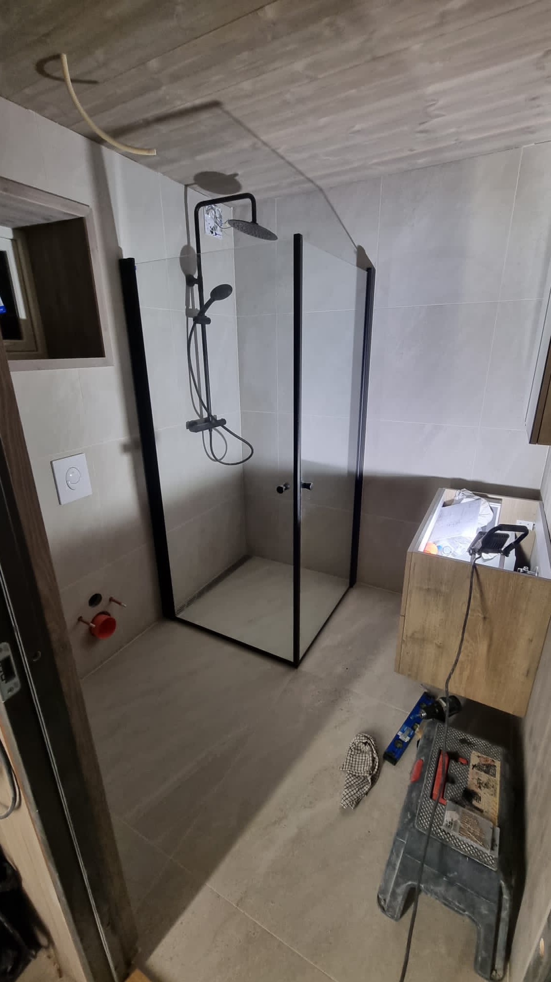 Nowoczesna kabina prysznicowa z czarnymi ramami i deszczownicą w łazience wyłożonej jasnymi płytkami, widoczne narzędzia i niedokończona szafka pod umywalkę.