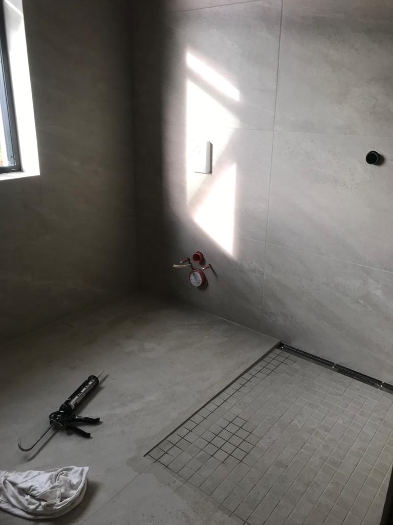 Szara łazienka w trakcie wykańczania, widoczny odpływ liniowy w podłodze, częściowo ułożone płytki, silikon w pistolecie, oraz wpadające światło przez okno.