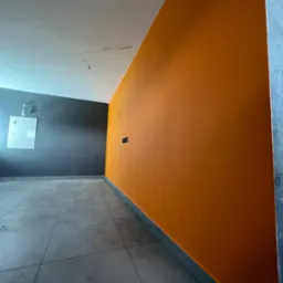 Świeżo pomalowane wnętrze z jedną ścianą w odcieniach pomarańczy, drugą w kolorze grafitowym, szara podłoga z płytek i wiszący kabel od oświetlenia.