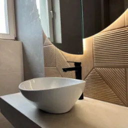 Nowoczesna łazienka z owalną białą umywalką nablatową, czarną baterią i dekoracyjną ścianą z podświetlanym lustrem o geometrycznym wzorze.